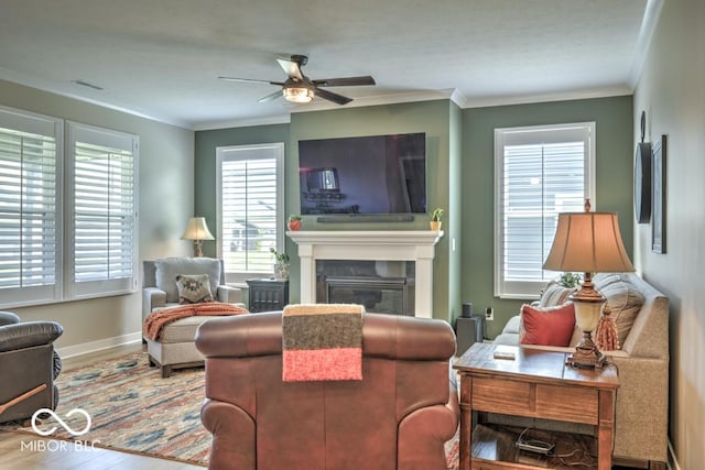 living area with wood finished floors, ceiling fan, ornamental molding, a glass covered fireplace, and baseboards