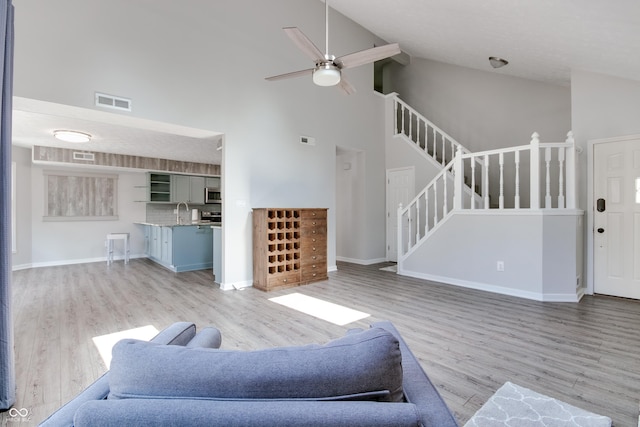 living area featuring high vaulted ceiling, stairs, light wood-style flooring, ceiling fan, and baseboards