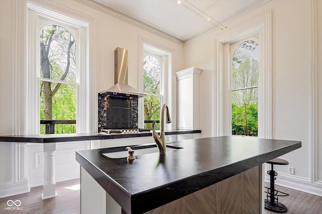 kitchen with wall chimney range hood, a sink, dark countertops, dark wood-style floors, and crown molding