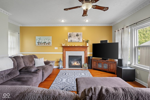 living area featuring ceiling fan, wood finished floors, a tile fireplace, ornamental molding, and baseboards