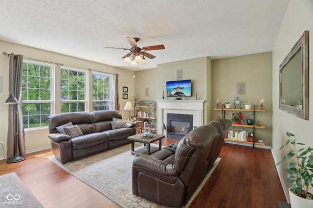 living room featuring ceiling fan, wood finished floors, a tiled fireplace, baseboards, and a textured ceiling