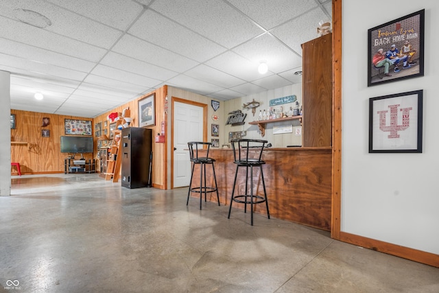 indoor dry bar featuring a paneled ceiling, baseboards, wood walls, and concrete flooring