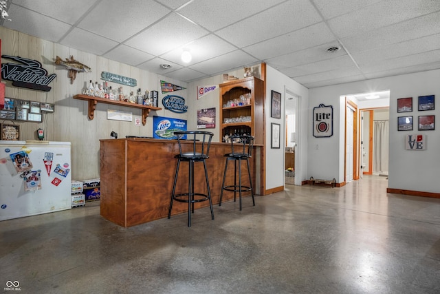 indoor dry bar with freestanding refrigerator, a drop ceiling, wooden walls, and baseboards