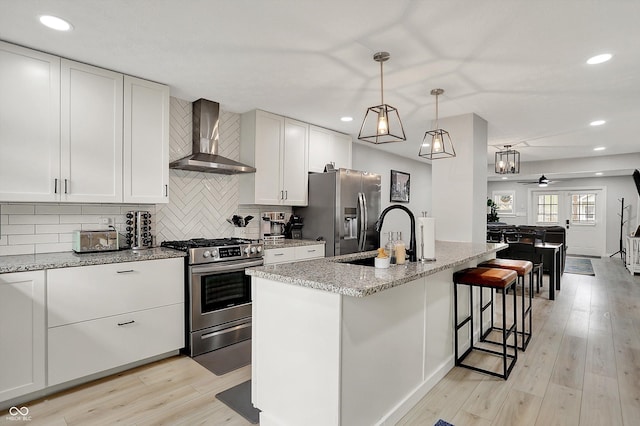 kitchen featuring stainless steel appliances, a sink, wall chimney exhaust hood, light stone counters, and ceiling fan
