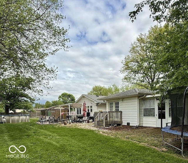 rear view of house with a trampoline, a deck, an outdoor pool, a yard, and a shingled roof