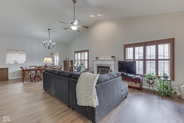 living room featuring a ceiling fan, light wood-style floors, a fireplace, high vaulted ceiling, and a chandelier
