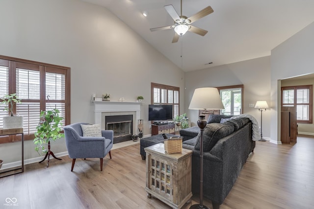 living room featuring high vaulted ceiling, a fireplace with flush hearth, a ceiling fan, light wood-style flooring, and baseboards
