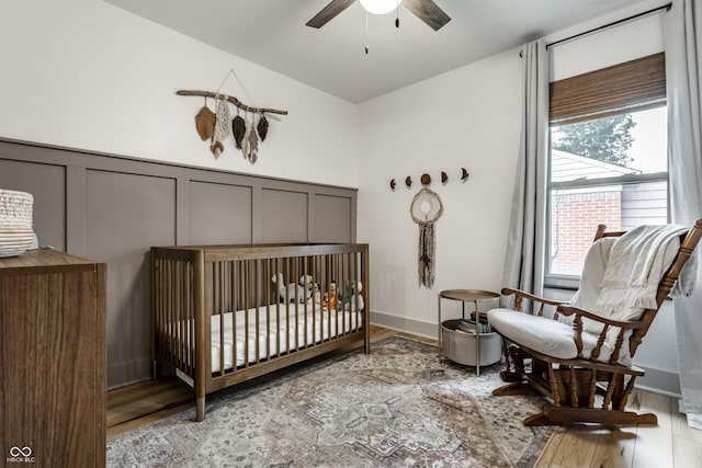 bedroom with a decorative wall, wood finished floors, a wainscoted wall, a ceiling fan, and a crib