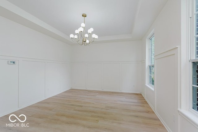 unfurnished dining area with a decorative wall, a chandelier, light wood-style flooring, a tray ceiling, and a wainscoted wall