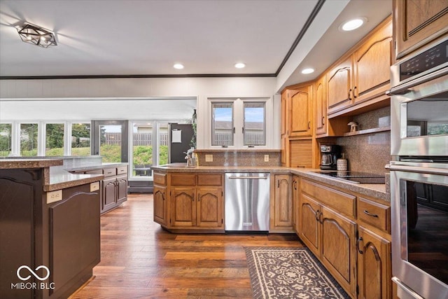 kitchen with appliances with stainless steel finishes, a peninsula, tasteful backsplash, dark wood-type flooring, and crown molding