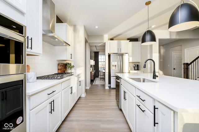 kitchen with a sink, wall chimney range hood, appliances with stainless steel finishes, light wood-style floors, and backsplash