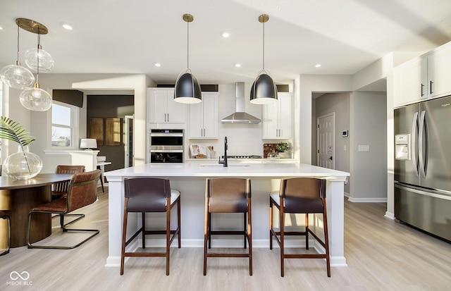 kitchen with stainless steel appliances, wall chimney range hood, light wood-style flooring, decorative backsplash, and light countertops