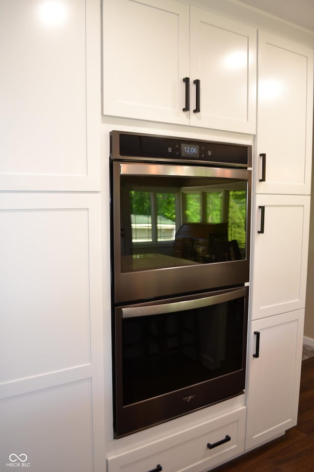 kitchen view of stainless steel double oven, white cabinets, and dark wood-style flooring