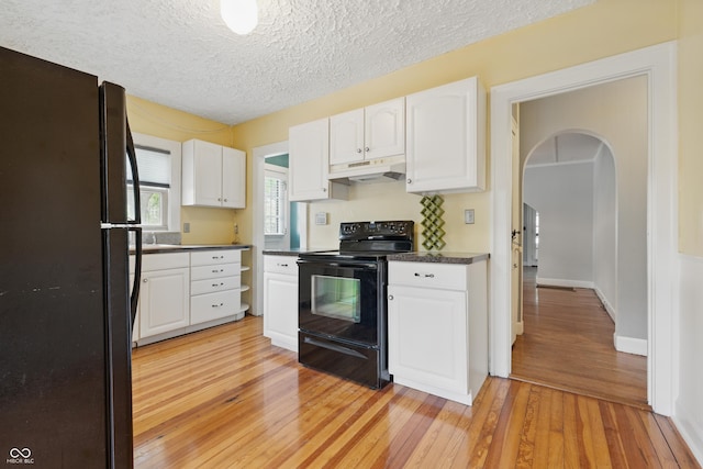 kitchen with black appliances, under cabinet range hood, light wood-type flooring, dark countertops, and arched walkways