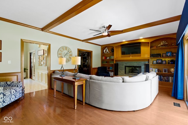 living room with wainscoting, a glass covered fireplace, a ceiling fan, wood finished floors, and crown molding
