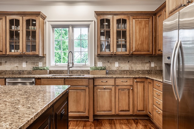 kitchen featuring stainless steel appliances, a sink, crown molding, light stone counters, and brown cabinets