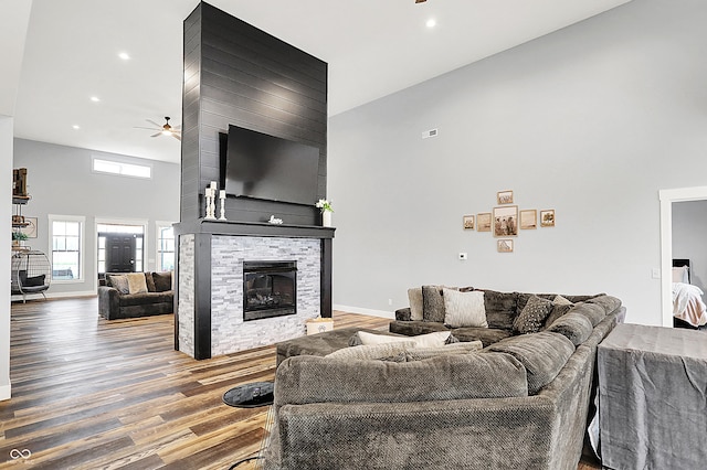 living area featuring a ceiling fan, a towering ceiling, wood finished floors, a stone fireplace, and recessed lighting