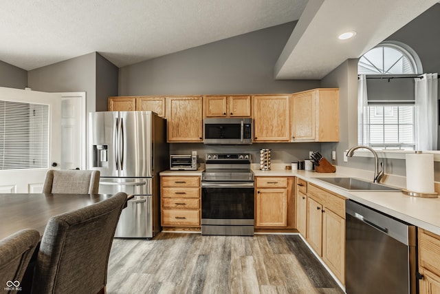 kitchen featuring stainless steel appliances, a sink, light brown cabinets, light wood-style floors, and lofted ceiling