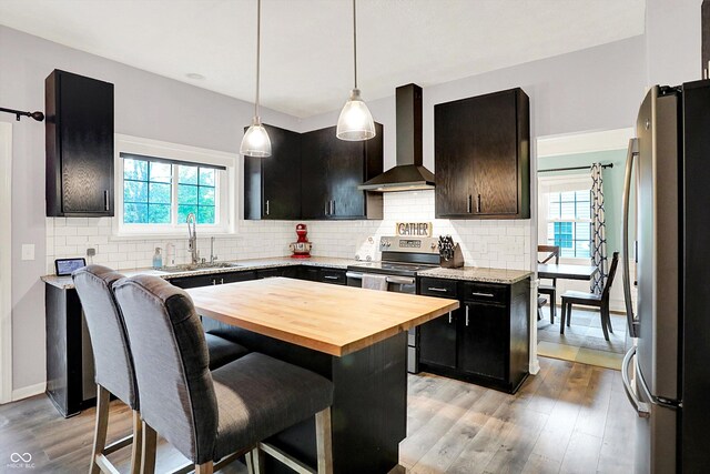 kitchen featuring wall chimney exhaust hood, stainless steel appliances, a sink, wood counters, and a breakfast bar area
