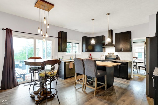 kitchen featuring wall chimney range hood, a center island, appliances with stainless steel finishes, backsplash, and wood counters