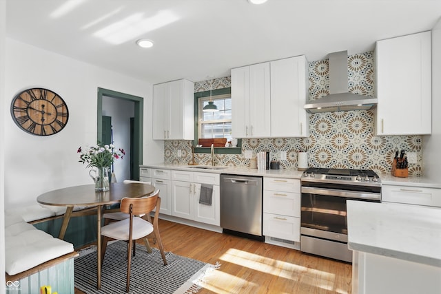 kitchen featuring stainless steel appliances, wall chimney exhaust hood, a sink, light wood-style floors, and tasteful backsplash