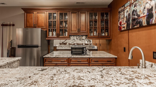 kitchen with freestanding refrigerator, brown cabinetry, light stone counters, glass insert cabinets, and a sink