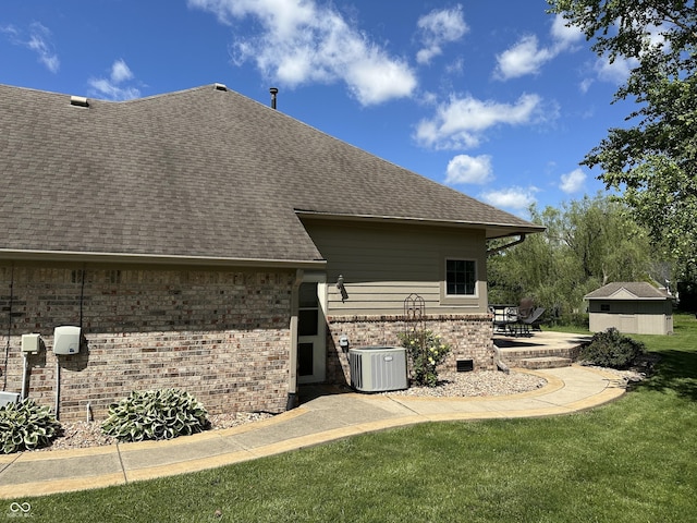 rear view of house with a shingled roof, cooling unit, a lawn, brick siding, and a patio