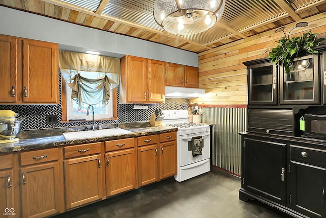 kitchen featuring white gas stove, a sink, wood walls, under cabinet range hood, and tasteful backsplash