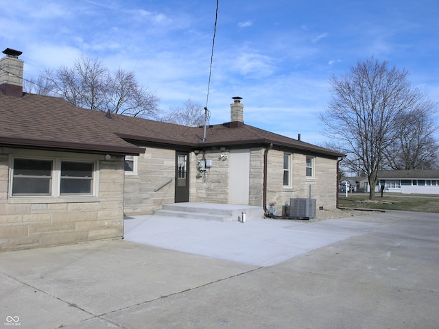 3456 S Sadlier Dr S, Indianapolis, IN 46239 - back of property with stone siding, a chimney, roof with shingles, and cooling unit