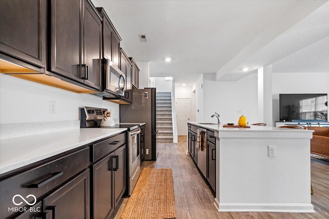 6507 W Whispering Way, Greenfield, IN 46140 - kitchen with stainless steel appliances, a sink, light wood-type flooring, dark brown cabinetry, and a kitchen island with sink