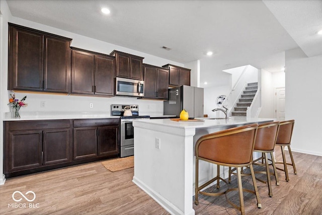 6507 W Whispering Way, Greenfield, IN 46140 - kitchen featuring stainless steel appliances, light wood-type flooring, an island with sink, dark brown cabinetry, and a breakfast bar area