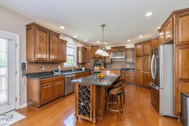 12772 Tram Ln, Carmel, IN 46032 - kitchen with appliances with stainless steel finishes, under cabinet range hood, a center island, light wood-type flooring, and brown cabinetry