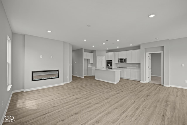 12500 Morning Ridge Way, Noblesville, IN 46060 - unfurnished living room featuring baseboards, light wood finished floors, recessed lighting, a glass covered fireplace, and a sink
