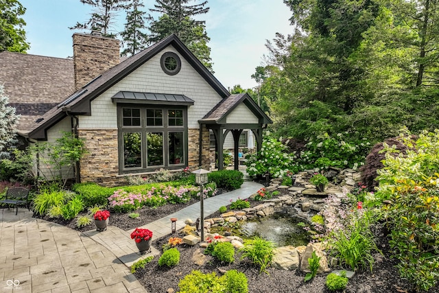 8651 Emerald Ln, Indianapolis, IN 46260 - view of front of house with stone siding, a chimney, a small pond, a standing seam roof, and a patio area
