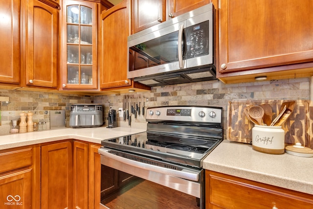 705 Ridge Gate Dr, Brownsburg, IN 46112 - kitchen with appliances with stainless steel finishes, tasteful backsplash, light countertops, brown cabinets, and glass insert cabinets