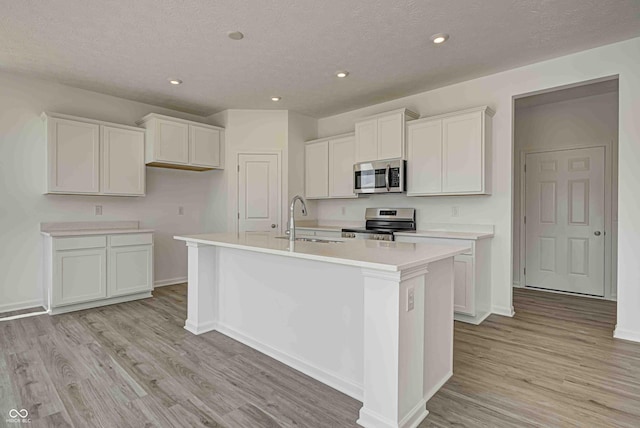 7501 Firecrest Ln, Camby, IN 46113 - kitchen with appliances with stainless steel finishes, a center island with sink, light wood finished floors, white cabinetry, and a textured ceiling