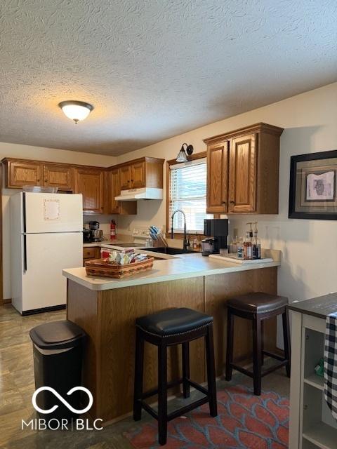 18 Hough St, Greenfield, IN 46140 - kitchen featuring freestanding refrigerator, under cabinet range hood, a peninsula, brown cabinetry, and a breakfast bar area