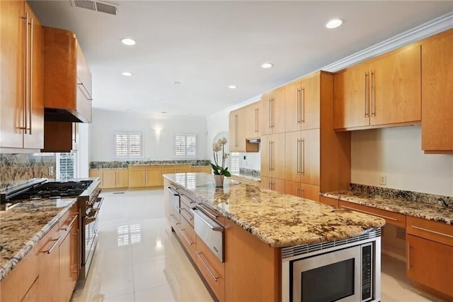 kitchen with appliances with stainless steel finishes, light tile flooring, a center island, light stone counters, and wall chimney exhaust hood