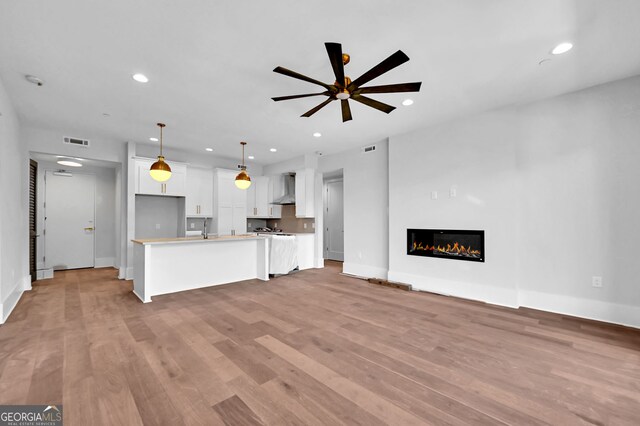 kitchen with ceiling fan, wall chimney exhaust hood, white cabinets, a kitchen island with sink, and light wood-type flooring