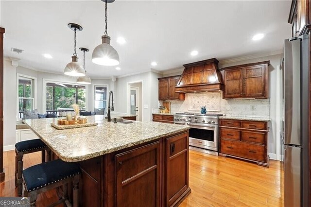 kitchen with appliances with stainless steel finishes, decorative light fixtures, light hardwood / wood-style floors, custom exhaust hood, and an island with sink