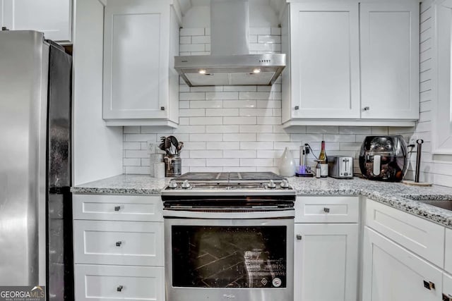 kitchen with tasteful backsplash, wall chimney range hood, stainless steel appliances, light stone countertops, and white cabinets