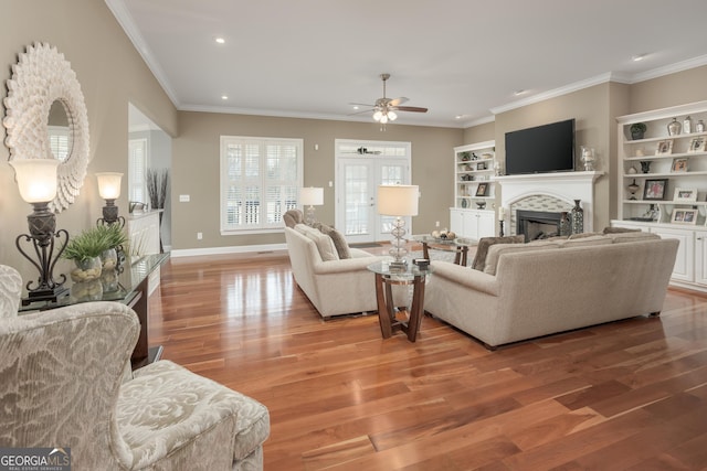 living room with ceiling fan, a fireplace, light hardwood / wood-style flooring, built in shelves, and crown molding