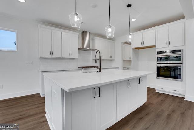 kitchen featuring wall chimney exhaust hood, a center island with sink, white cabinetry, and double oven