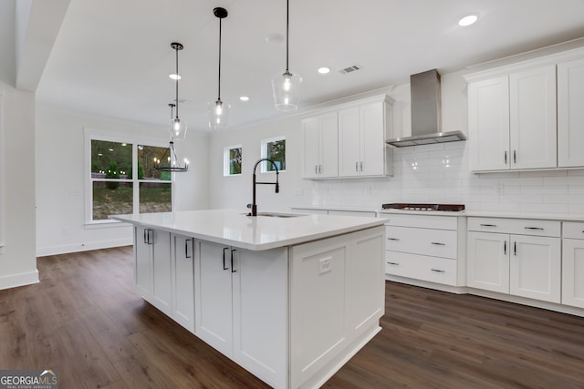 kitchen with a center island with sink, wall chimney range hood, hanging light fixtures, sink, and white cabinets