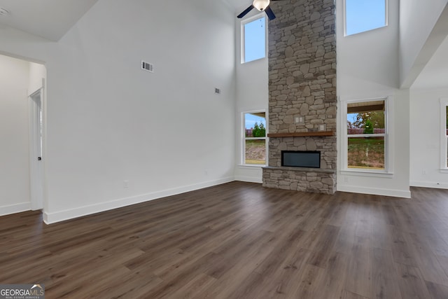 unfurnished living room with ceiling fan, a towering ceiling, a stone fireplace, and dark wood-type flooring