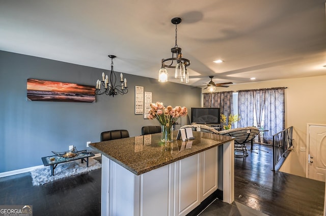 kitchen with hanging light fixtures, ceiling fan with notable chandelier, dark wood-type flooring, dark stone counters, and white cabinetry