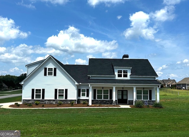 view of front facade featuring roof with shingles, a chimney, covered porch, a front yard, and stone siding