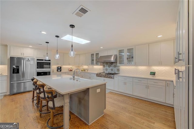 kitchen featuring a skylight, exhaust hood, appliances with stainless steel finishes, a center island with sink, and white cabinets