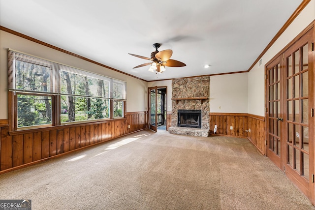 unfurnished living room with light colored carpet, a stone fireplace, wooden walls, ceiling fan, and ornamental molding