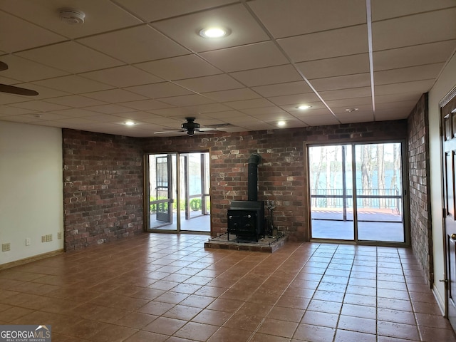 unfurnished living room with brick wall, ceiling fan, a drop ceiling, dark tile flooring, and a wood stove